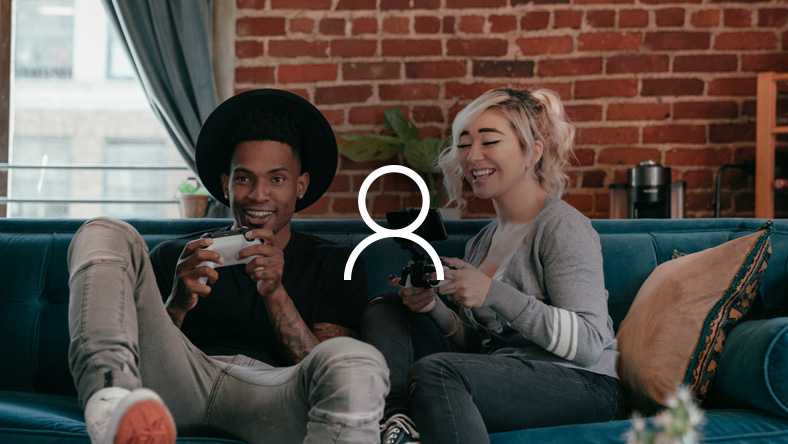 A man in a black hat and a blonde woman are smiling together as they game on a couch with Xbox controllers.